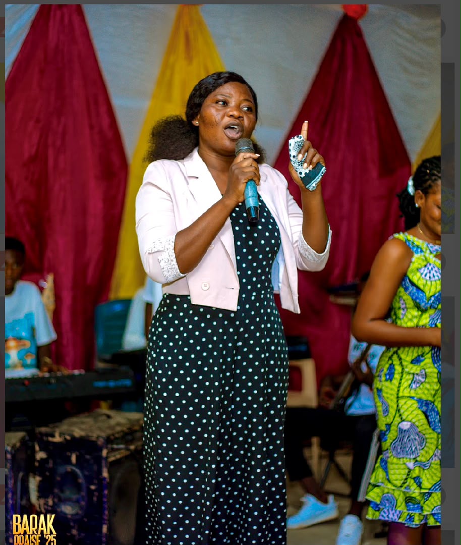 Minister Vida with a hand raised, singing in front of red and gold stage drapes at Barak Praise 2025.