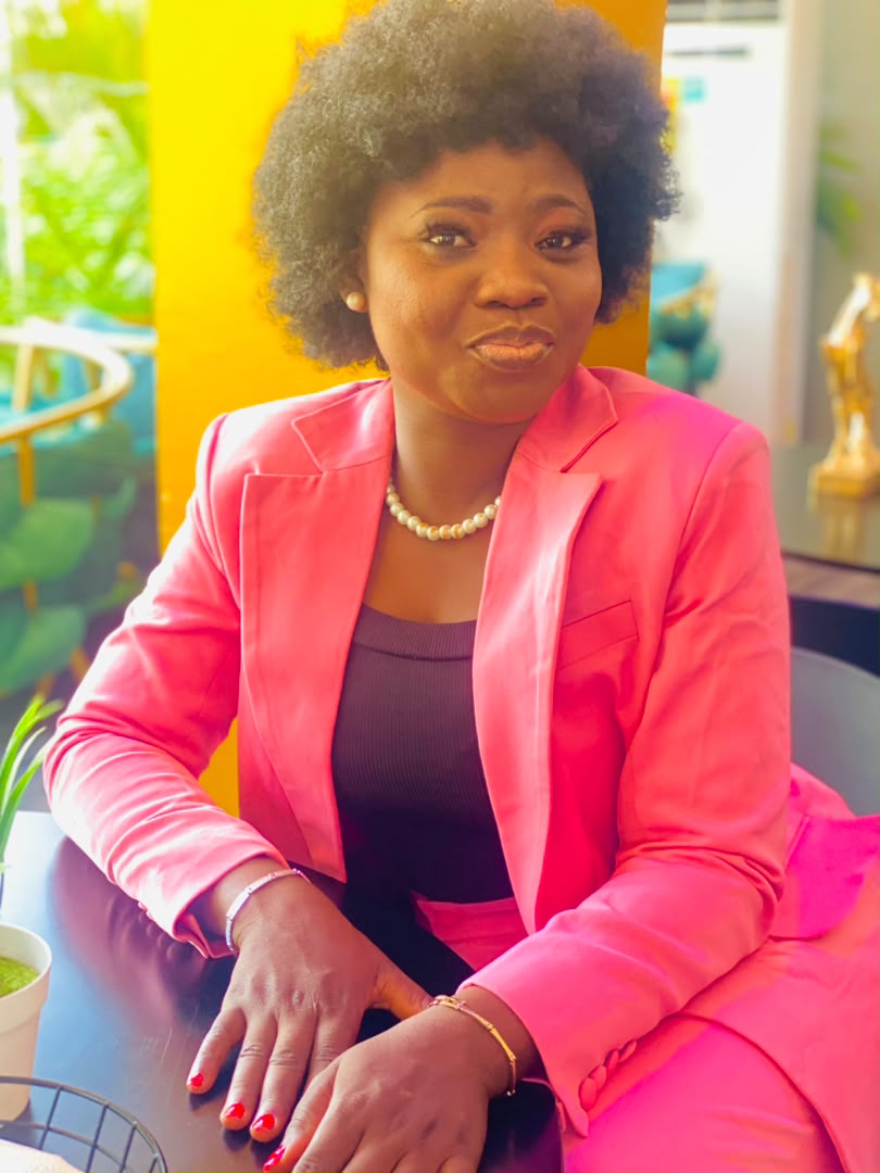 Minister Vida seated in a pink suit against a yellow wall.